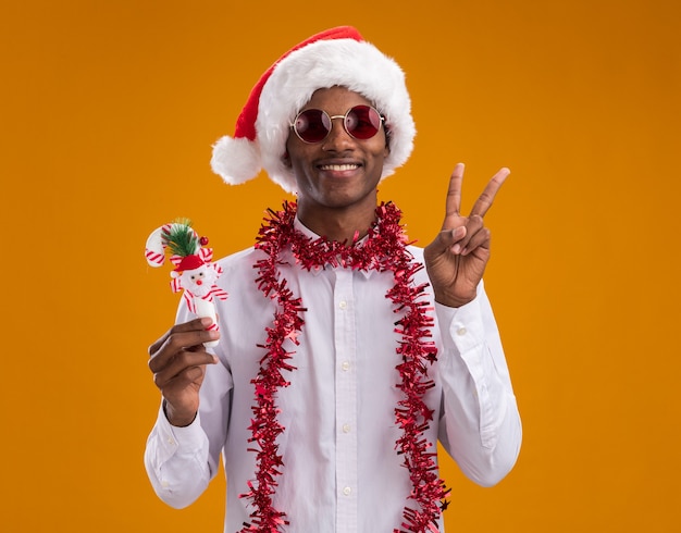 Souriant jeune homme afro-américain portant bonnet de Noel et lunettes avec guirlande de guirlandes autour du cou tenant l'ornement de canne à sucre faisant signe de paix isolé sur mur orange