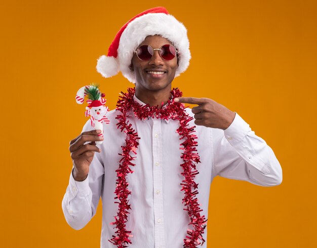 Souriant jeune homme afro-américain portant bonnet de Noel et lunettes avec guirlande de guirlandes autour du cou tenant et pointant sur l'ornement de canne à sucre en regardant la caméra isolée sur fond orange