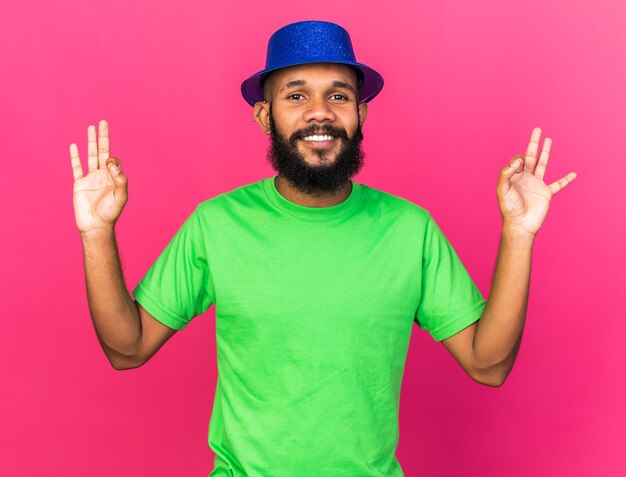 Photo gratuite souriant jeune homme afro-américain portant un chapeau de fête montrant un geste correct isolé sur un mur rose