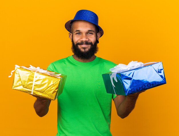 Souriant jeune homme afro-américain portant un chapeau de fête tenant des coffrets cadeaux isolés sur un mur orange