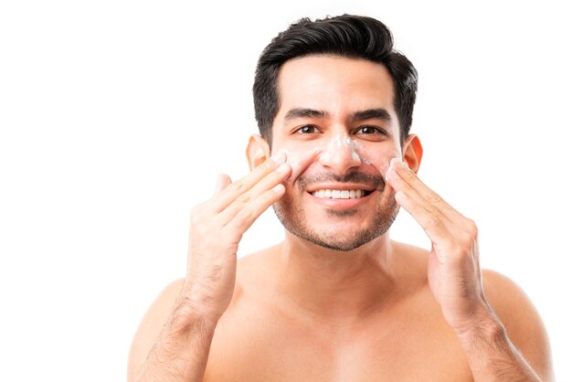 Souriant jeune homme appliquant une crème anti-âge sur les joues tout en établissant un contact visuel sur fond blanc