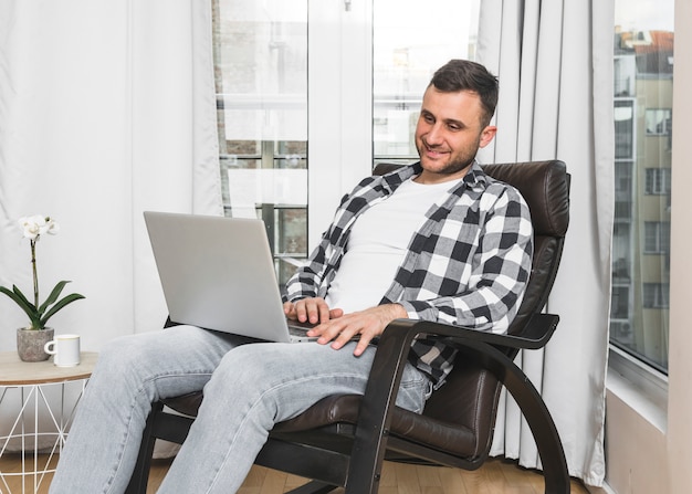 Souriant jeune homme assis sur une chaise à l&#39;aide de téléphone portable à la maison