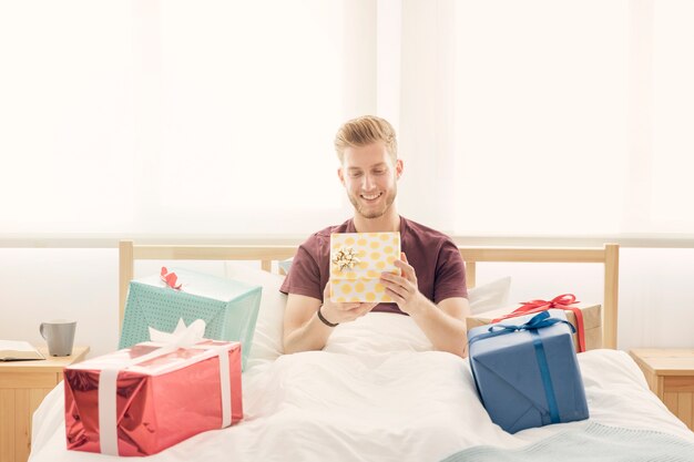 Photo gratuite souriant jeune homme assis sur un lit entouré de cadeaux colorés
