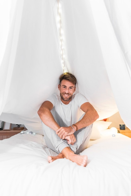 Souriant jeune homme assis sur le lit sous le rideau blanc