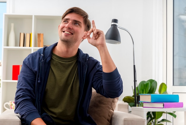 Souriant jeune homme beau blond est assis sur un fauteuil pointant vers le haut en regardant le côté à l'intérieur du salon