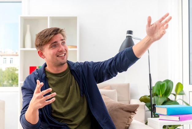 Souriant jeune homme beau blond est assis sur un fauteuil à la recherche et pointant sur le côté avec deux mains