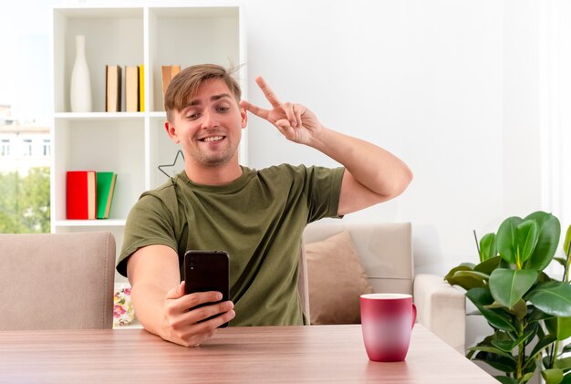 Souriant jeune homme beau blond est assis à table avec tasse tenant et regardant le téléphone gestes signe de la main de la victoire à l'intérieur du salon