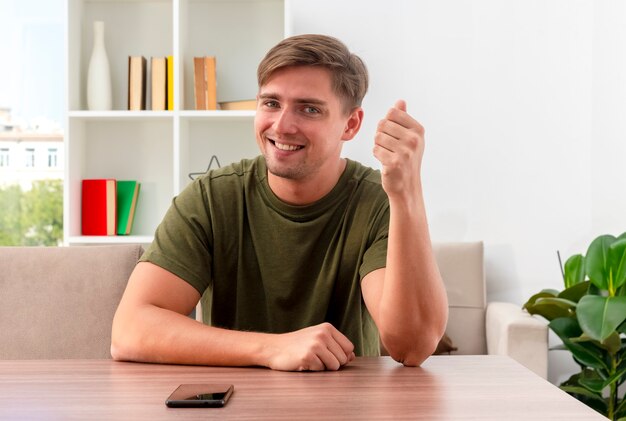 Souriant jeune homme beau blond est assis à table avec téléphone en gardant le poing levé et regardant la caméra à l'intérieur du salon