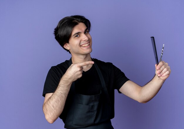 Souriant jeune homme beau coiffeur en tenue uniforme et points au peigne avec des ciseaux isolé sur bleu