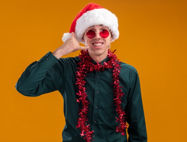 Souriant jeune homme blond portant bonnet de Noel et lunettes avec guirlande de guirlandes autour du cou regardant la caméra faisant signe d'appel isolé sur fond orange