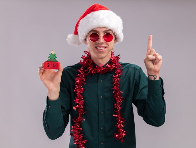 Souriant jeune homme blond portant un bonnet de Noel et des lunettes avec une guirlande de guirlandes autour du cou tenant un jouet d'arbre de Noël avec une date regardant la caméra pointant vers le haut isolé sur fond blanc