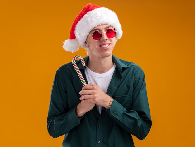 Photo gratuite souriant jeune homme blond portant bonnet de noel et lunettes tenant une canne douce de noël regardant la caméra isolée sur fond orange
