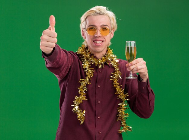 Souriant jeune homme blond portant des lunettes avec guirlande de guirlandes autour du cou tenant un verre de champagne montrant le pouce vers le haut isolé sur mur vert