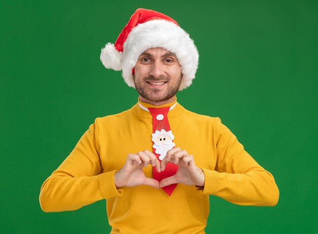 Souriant jeune homme caucasien portant chapeau de Noël et cravate faisant signe de coeur regardant la caméra isolée sur fond vert