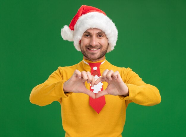 Souriant Jeune Homme Caucasien Portant Chapeau De Noël Et Cravate Regardant La Caméra Faisant Signe De Coeur Isolé Sur Fond Vert