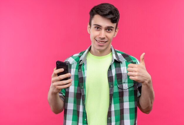 Souriant jeune homme caucasien portant une chemise verte tenant le téléphone son pouce vers le haut sur fond rose isolé