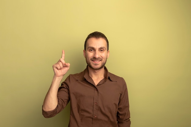 Souriant jeune homme caucasien regardant la caméra pointant vers le haut isolé sur fond vert olive avec espace copie