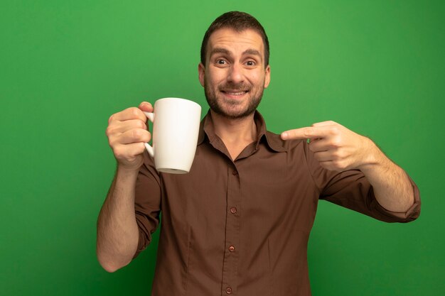 Souriant jeune homme caucasien tenant et pointant sur une tasse de thé en regardant la caméra isolée sur fond vert