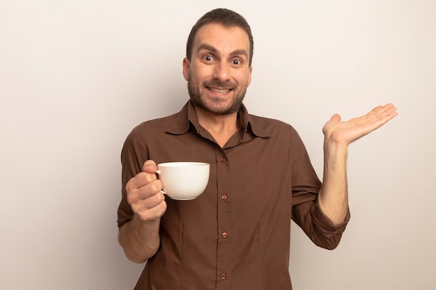 Souriant jeune homme caucasien tenant une tasse de thé regardant la caméra montrant la main vide isolé sur fond blanc avec espace copie