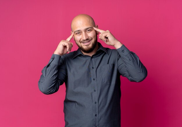 Souriant jeune homme de centre d'appels chauve mettant les doigts sur les tempes isolées sur mur cramoisi