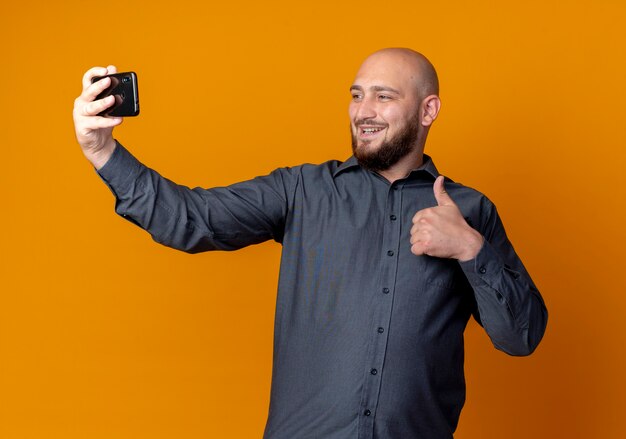 Souriant jeune homme de centre d'appels chauve montrant le pouce vers le haut et prenant selfie isolé sur mur orange