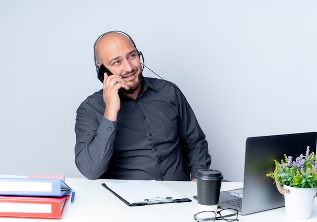 Souriant jeune homme de centre d'appels chauve portant un casque assis au bureau avec des outils de travail à côté et parler au téléphone isolé sur un mur blanc