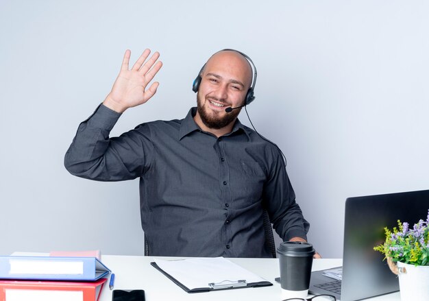 Souriant jeune homme de centre d'appels chauve portant un casque assis au bureau avec des outils de travail gestes salut à l'avant isolé sur un mur blanc