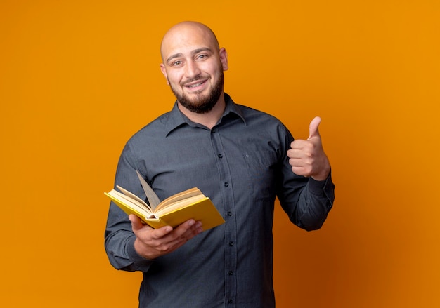 Souriant jeune homme de centre d'appels chauve tenant un livre montrant le pouce vers le haut isolé sur un mur orange