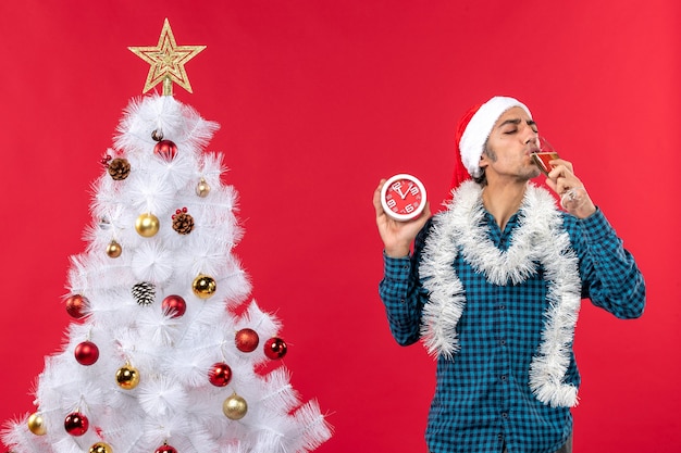 Souriant jeune homme avec chapeau de père Noël et boire un verre de vin et tenant une horloge debout près de l'arbre de Noël sur le rouge