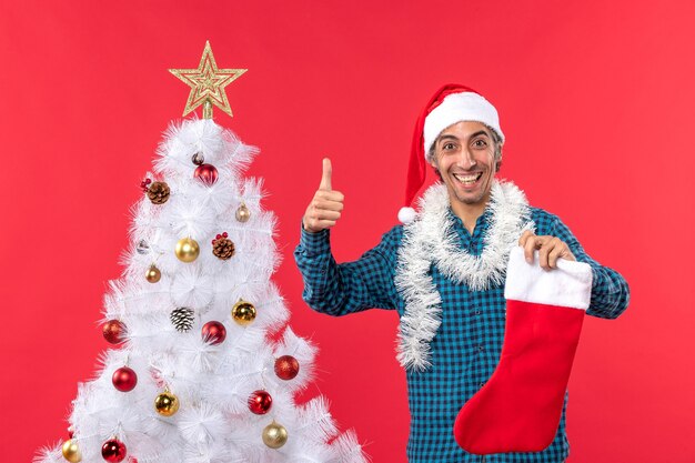 Souriant jeune homme avec chapeau de père Noël dans une chemise bleue dépouillée et tenant la chaussette de Noël faisant le geste ok