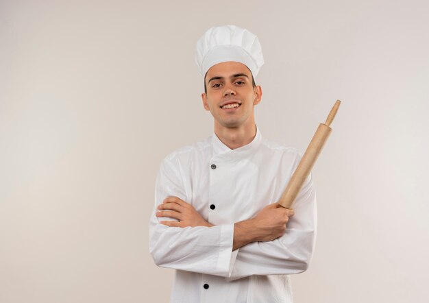 Souriant jeune homme cuisinier portant l'uniforme de chef croisant les mains tenant le rouleau à pâtisserie avec copie espace