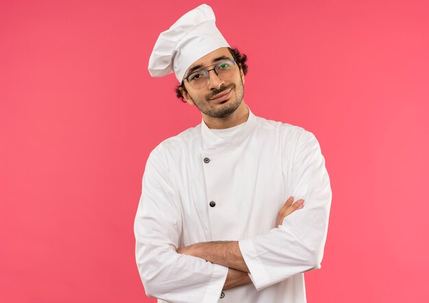 Souriant jeune homme cuisinier portant l'uniforme de chef et des lunettes croisant les mains sur le rose