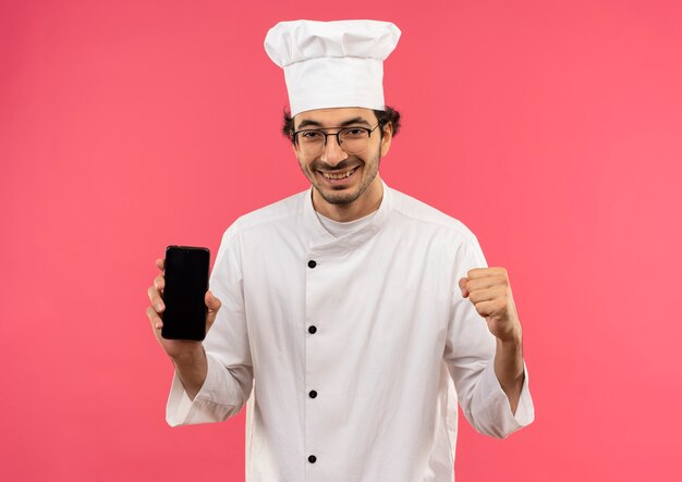 Souriant jeune homme cuisinier portant l'uniforme de chef et des lunettes tenant le téléphone et montrant oui geste isolé sur mur rose