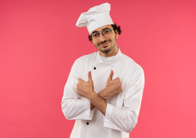 Souriant jeune homme cuisinier portant l'uniforme de chef et des lunettes traversant les mains ses pouces vers le haut isolé sur mur rose