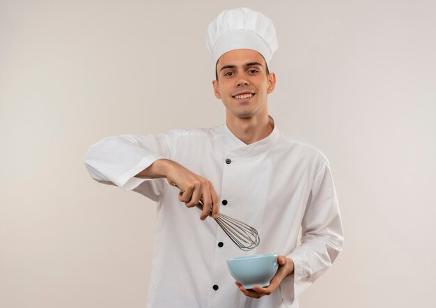 Souriant jeune homme cuisinier portant l'uniforme de chef tenant un fouet avec un bol