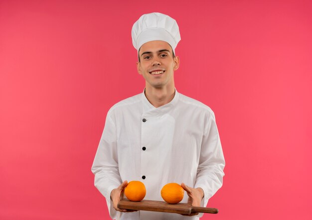 Souriant jeune homme cuisinier portant l'uniforme de chef tenant orange sur une planche à découper avec espace copie