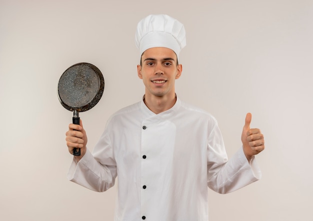 Souriant jeune homme cuisinier portant l'uniforme de chef tenant une poêle à frire son pouce vers le haut