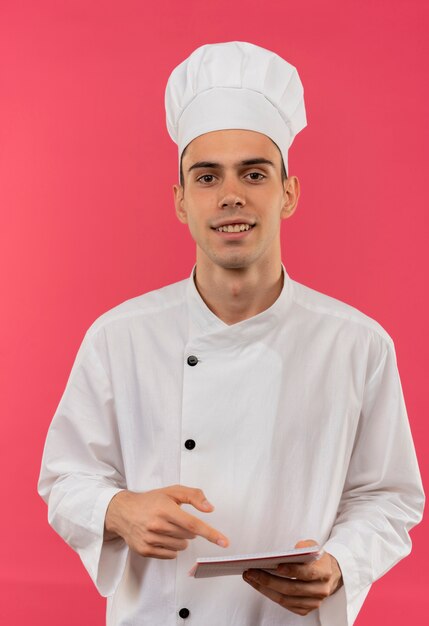 Souriant jeune homme cuisinier portant un uniforme de chef tenant et pointe vers le cahier