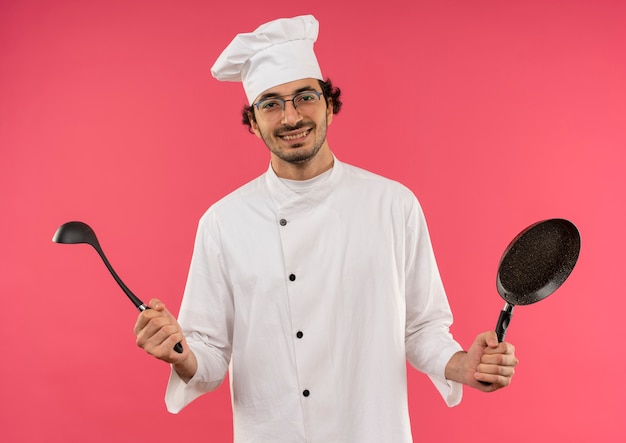 Souriant jeune homme cuisinier portant l'uniforme de chef et des verres tenant une poêle avec une louche sur rose