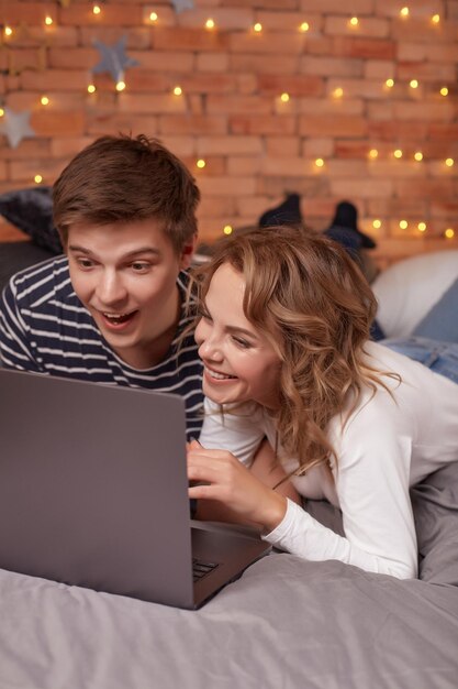 Souriant jeune homme et femme allongé dans son lit et regardant quelque chose sur l'ordinateur portable. Ils sont heureux et adorables