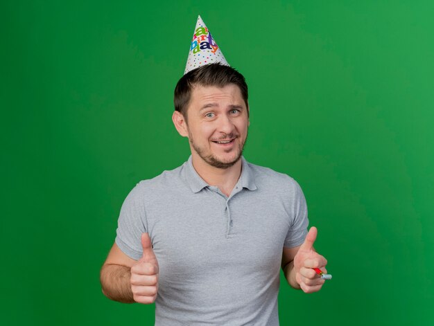 Souriant jeune homme de fête portant une casquette d'anniversaire tenant un sifflet montrant les pouces vers le haut isolé sur vert