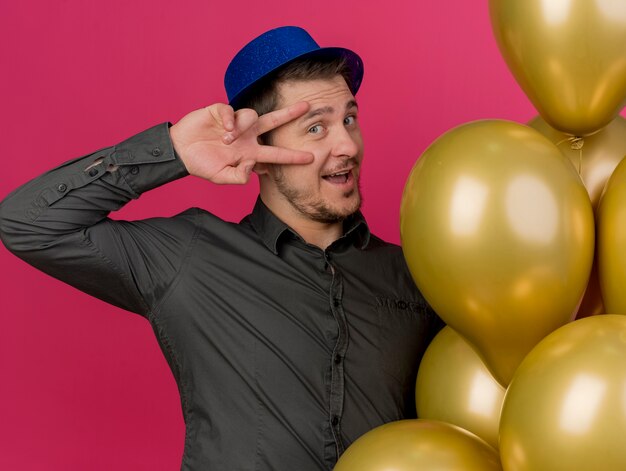 Souriant jeune homme de fête portant un chapeau bleu debout à côté des ballons montrant le geste de paix isolé sur rose