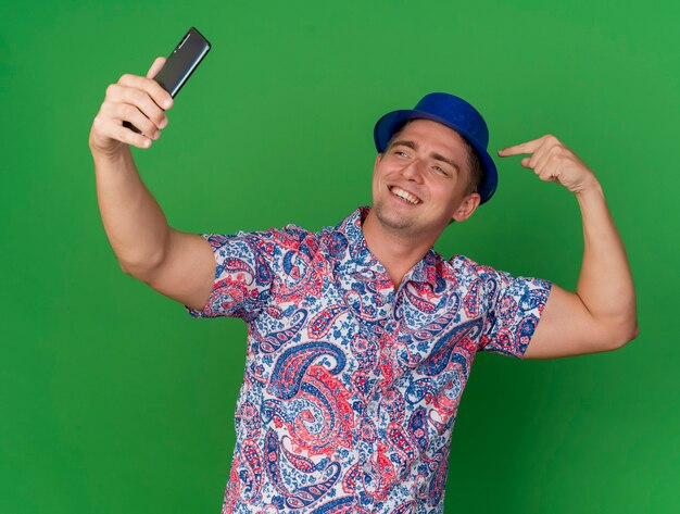 Souriant jeune homme de fête portant un chapeau bleu prend un selfie et se montre isolé sur fond vert