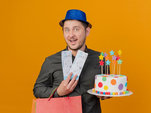 Souriant jeune homme de fête portant un chapeau bleu tenant un gâteau avec un sac-cadeau et des billets isolés sur orange