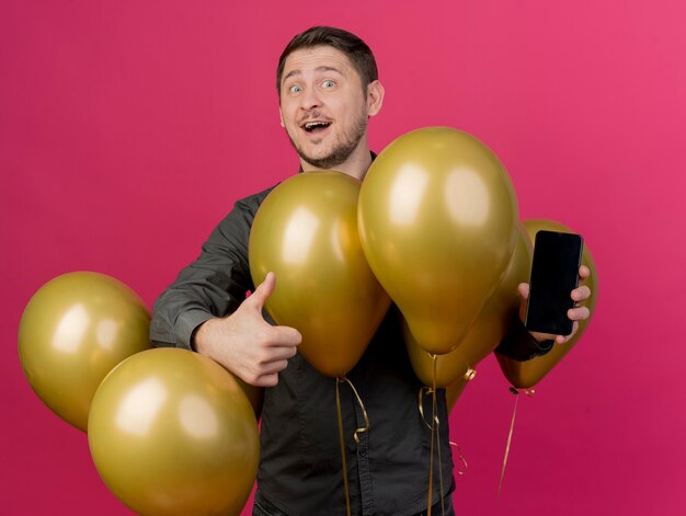 Souriant jeune homme de fête vêtu d'une chemise noire debout parmi les ballons tenant le téléphone montrant le pouce vers le haut isolé sur rose