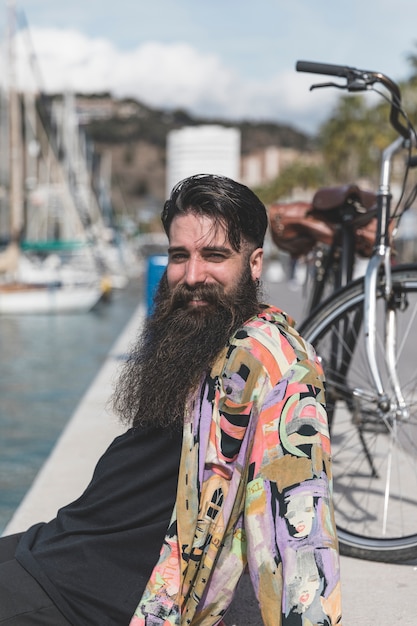 Photo gratuite souriant jeune homme avec une longue barbe assis sur la côte en regardant la caméra