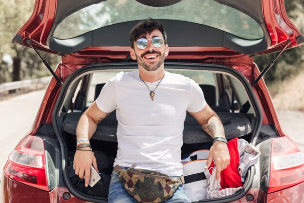 Souriant jeune homme avec des lunettes, s&#39;appuyant sur un coffre de voiture ouverte