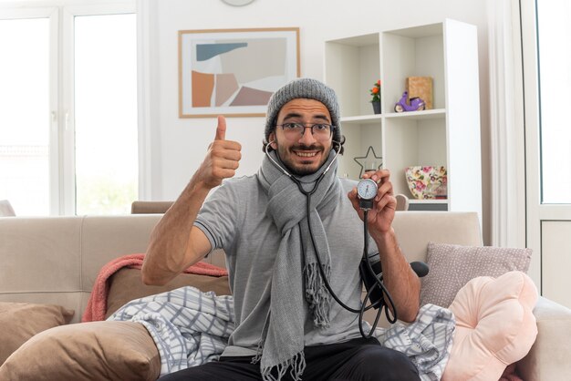 souriant jeune homme malade dans des lunettes optiques avec un foulard autour du cou portant un chapeau d'hiver mesurant la pression avec un sphygmomanomètre et levant le pouce assis sur un canapé dans le salon