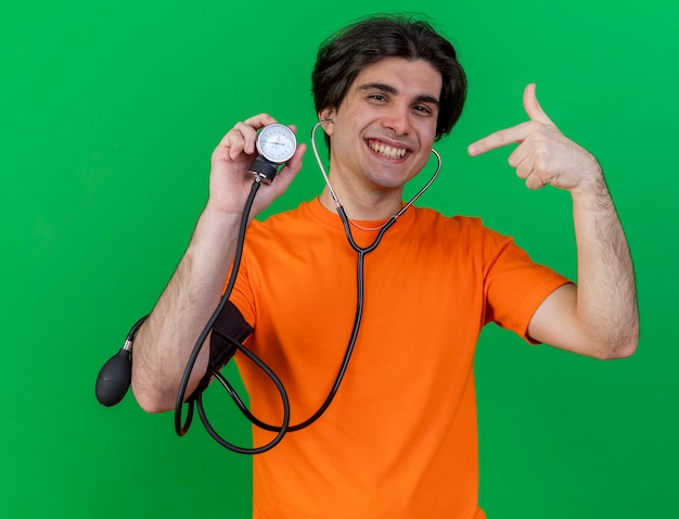 Souriant jeune homme malade portant stéthoscope tenant et points au sphygmomanomètre isolé sur vert