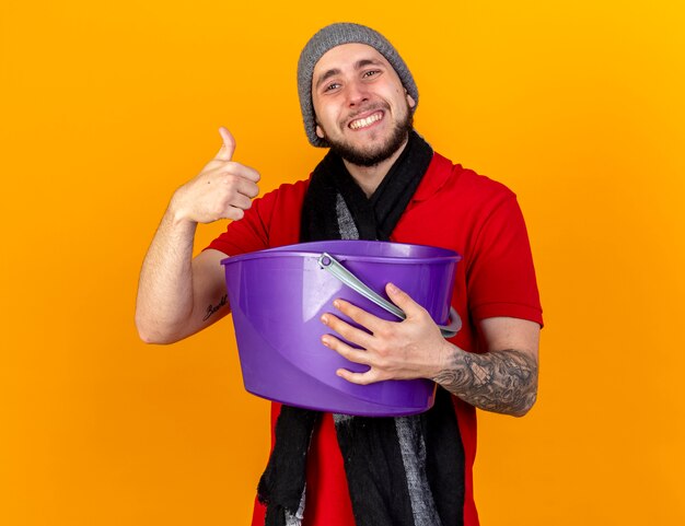 Souriant jeune homme malade de race blanche portant un chapeau d'hiver et une écharpe détient un seau en plastique et les pouces vers le haut isolé sur un mur orange avec copie espace